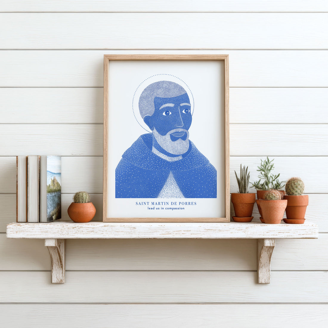 Blue St. Martin de Porres print on a white shelf beside books and small plants.
