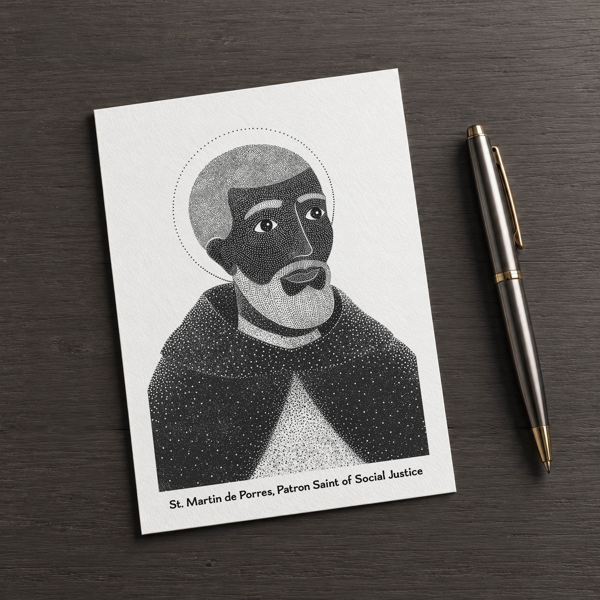 Black St Martin de Porres card on a dark table beside a pen