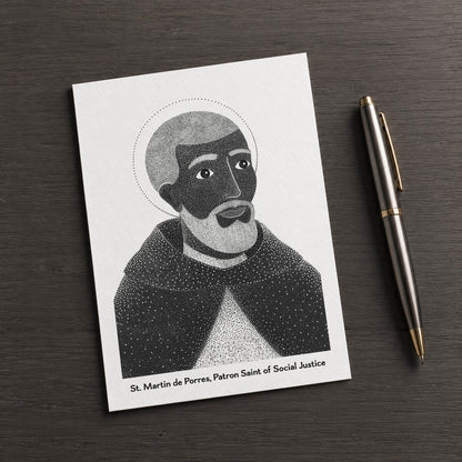 Black St Martin de Porres card on a dark table beside a pen