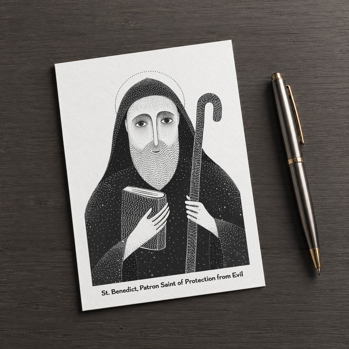 Black St Benedict card on a dark table beside a pen