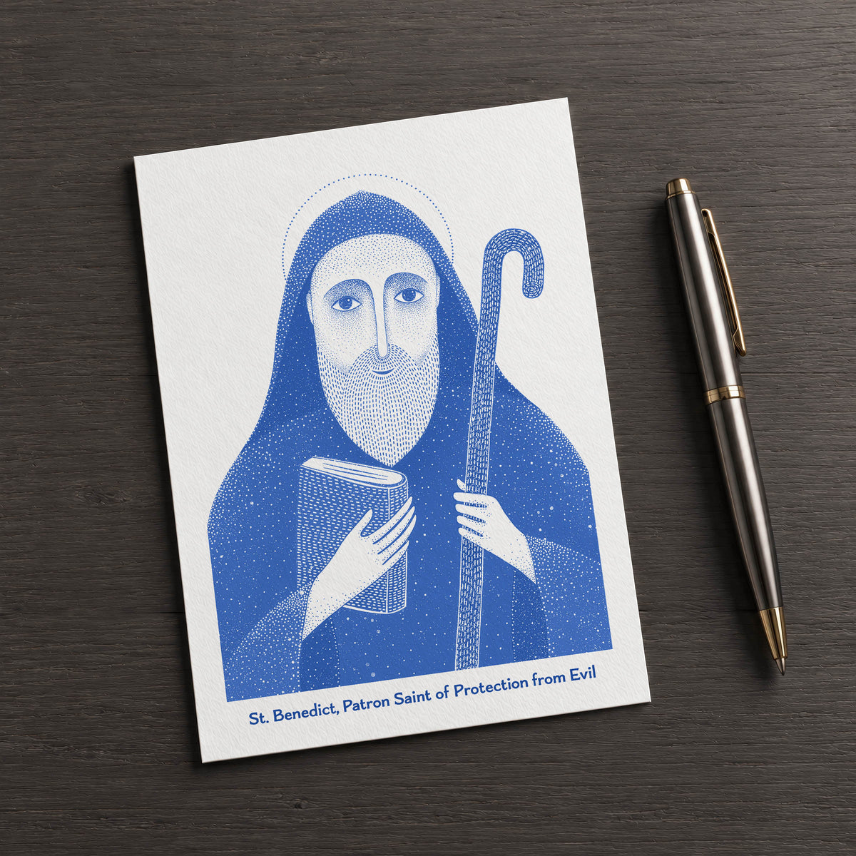 Blue St Benedict card on a dark table beside a pen