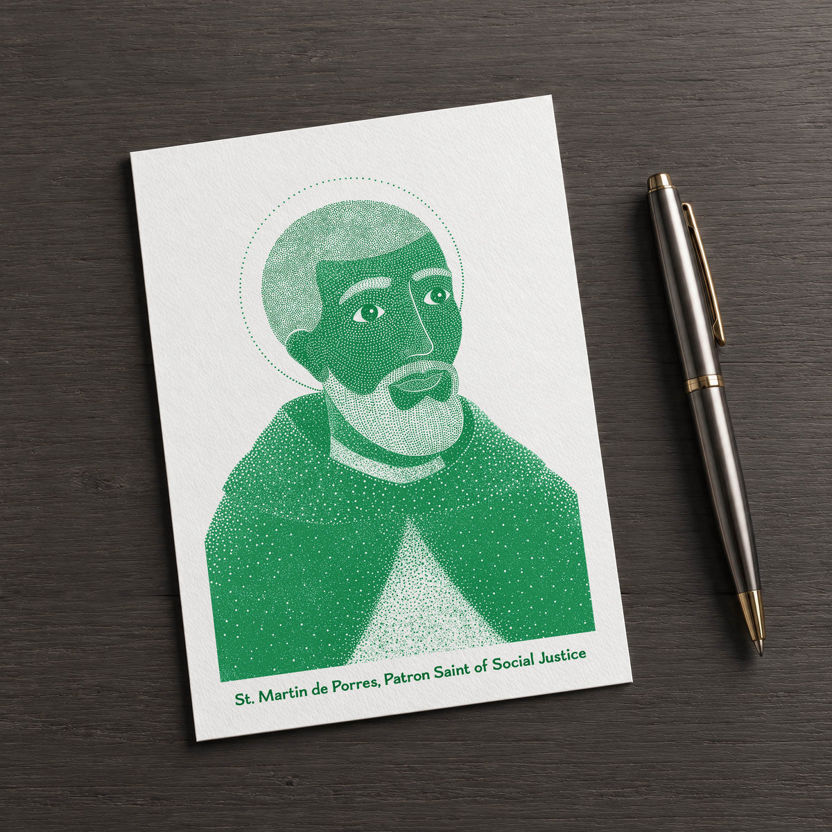 Green St Martin de Porres card on a dark table beside a pen
