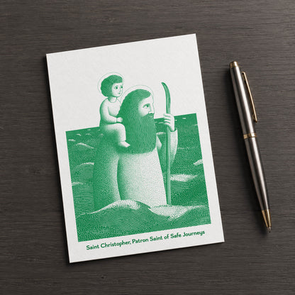 Green St Christopher card on a dark table beside a pen