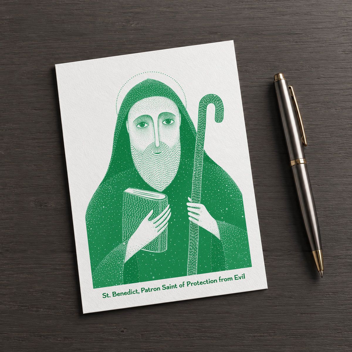 Green St Benedict card on a dark table beside a pen