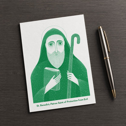 Green St Benedict card on a dark table beside a pen
