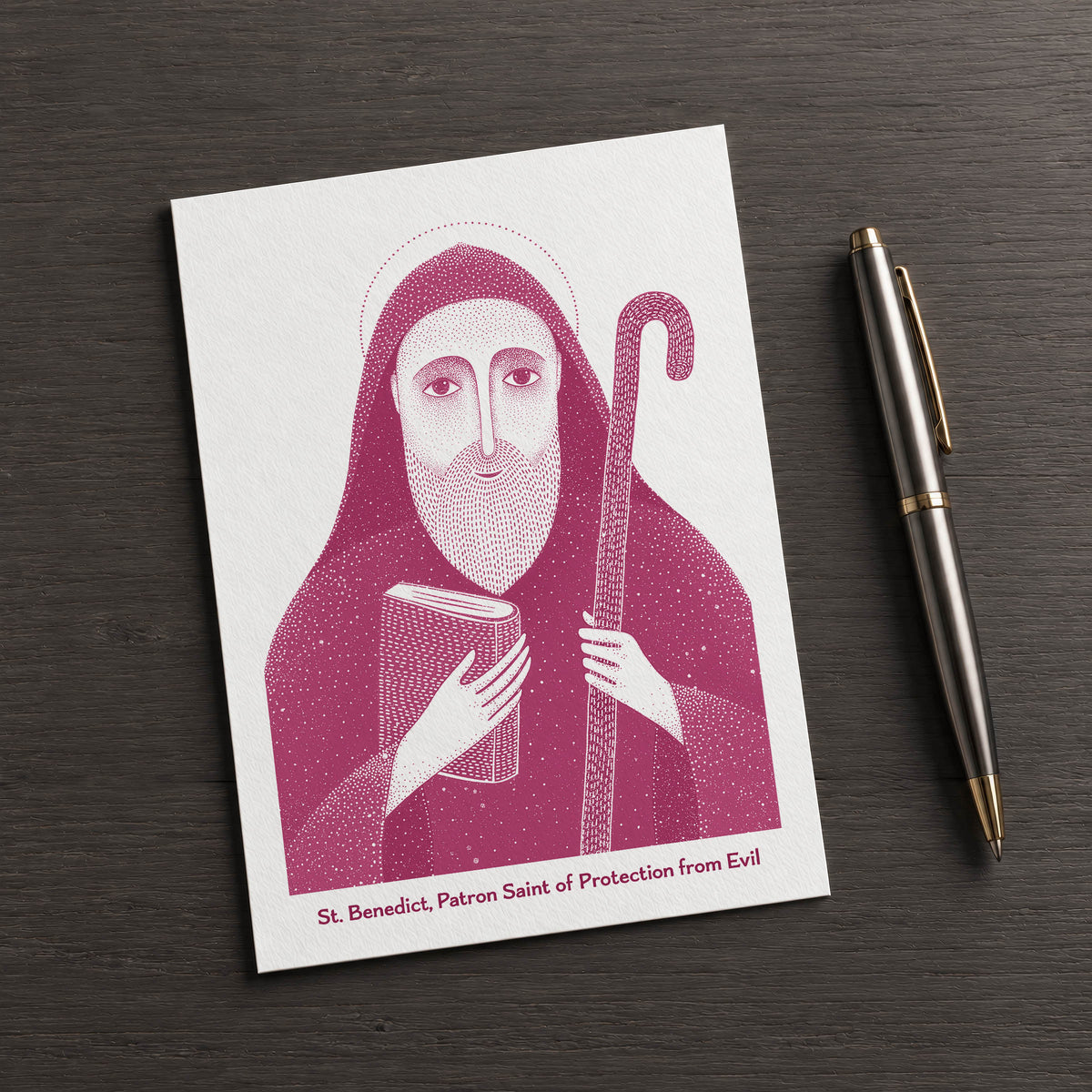 Pink St Benedict card on a dark table beside a pen