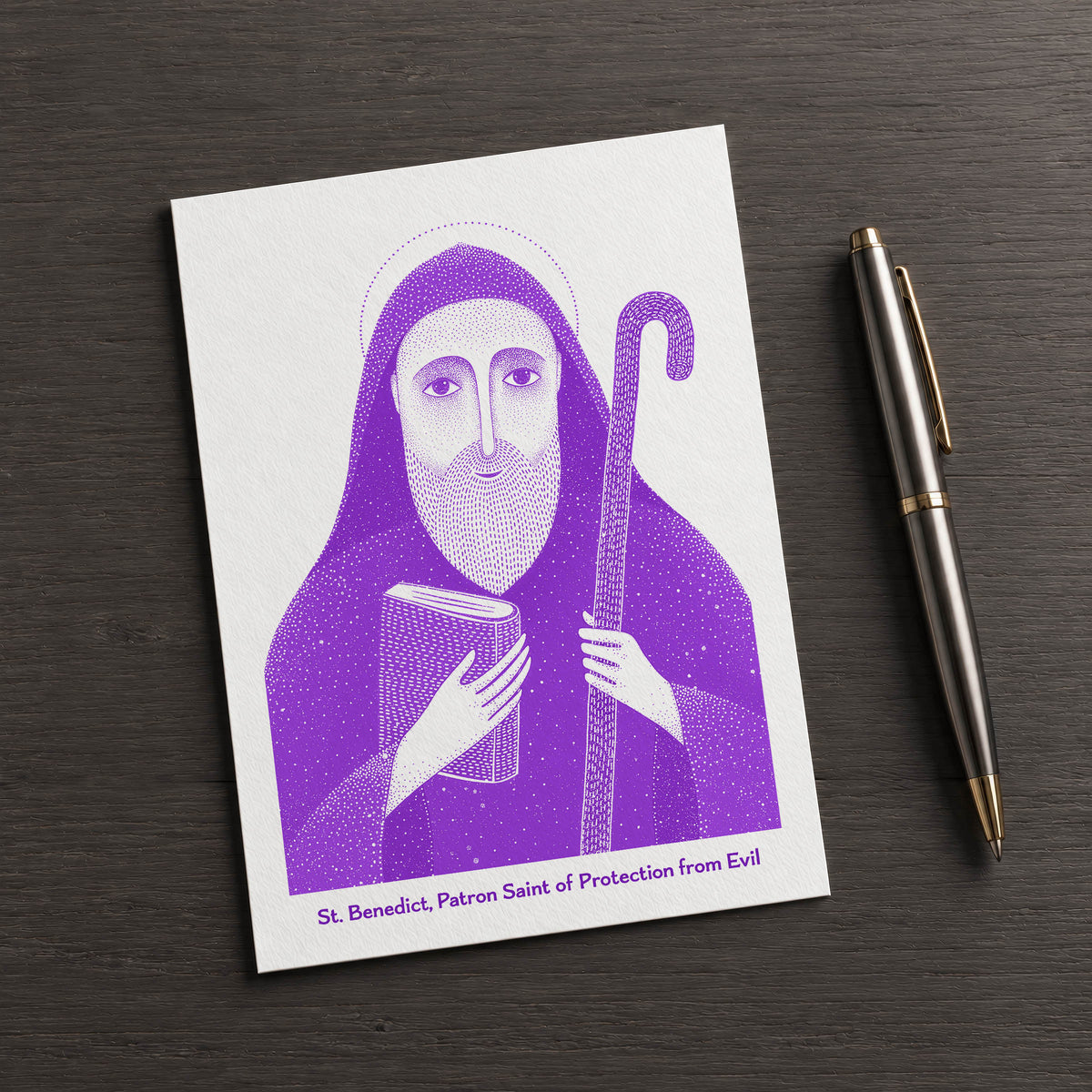 Purple St Benedict card on a dark table beside a pen