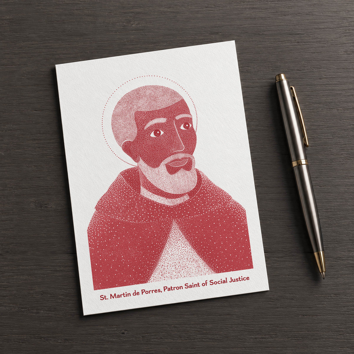 Red St Martin de Porres card on a dark table beside a pen