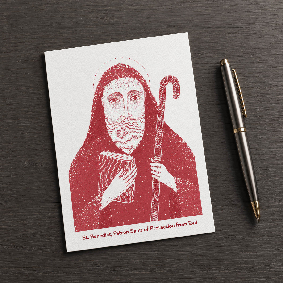 Red St Benedict card on a dark table beside a pen