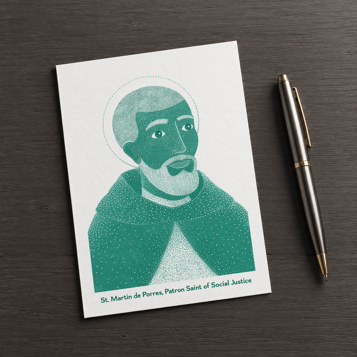 Teal St Martin de Porres card on a dark table beside a pen