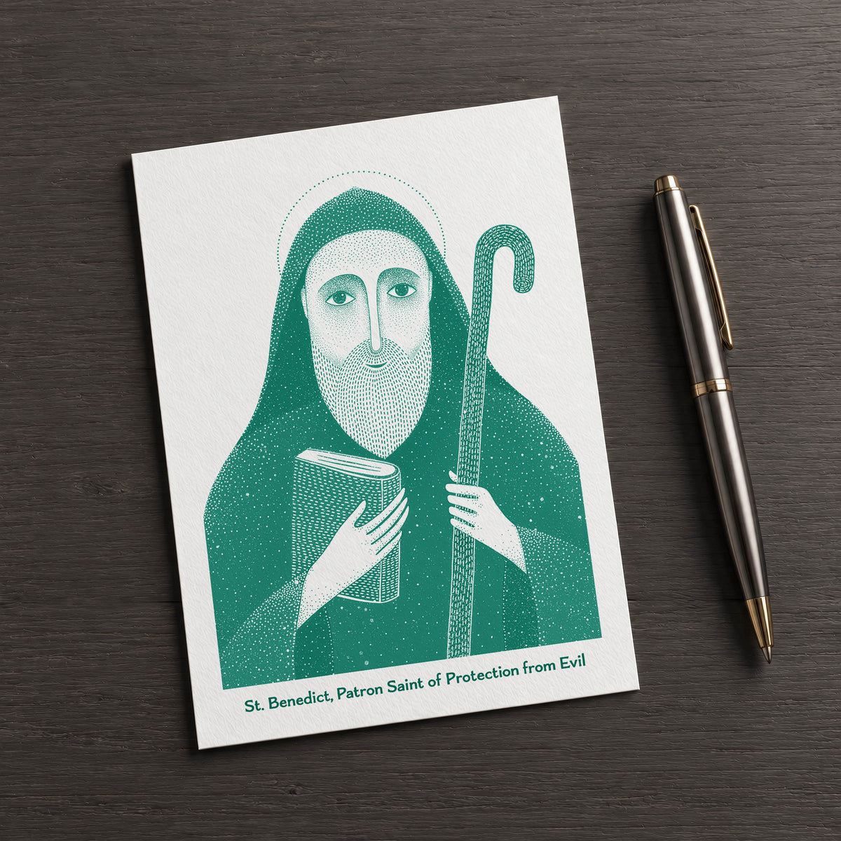 Teal St Benedict card on a dark table beside a pen