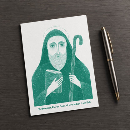Teal St Benedict card on a dark table beside a pen