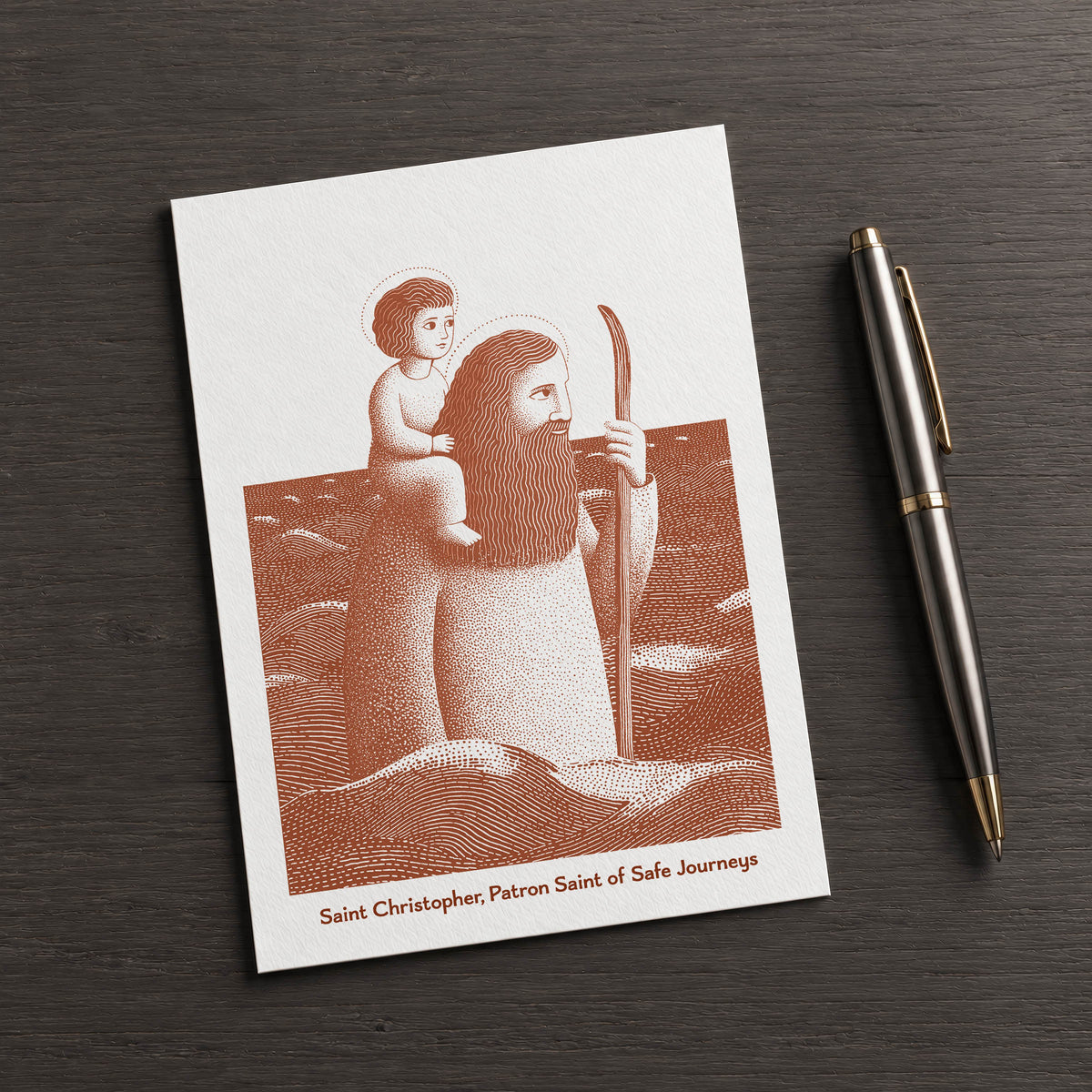 Terracotta St Christopher card on a dark table beside a pen